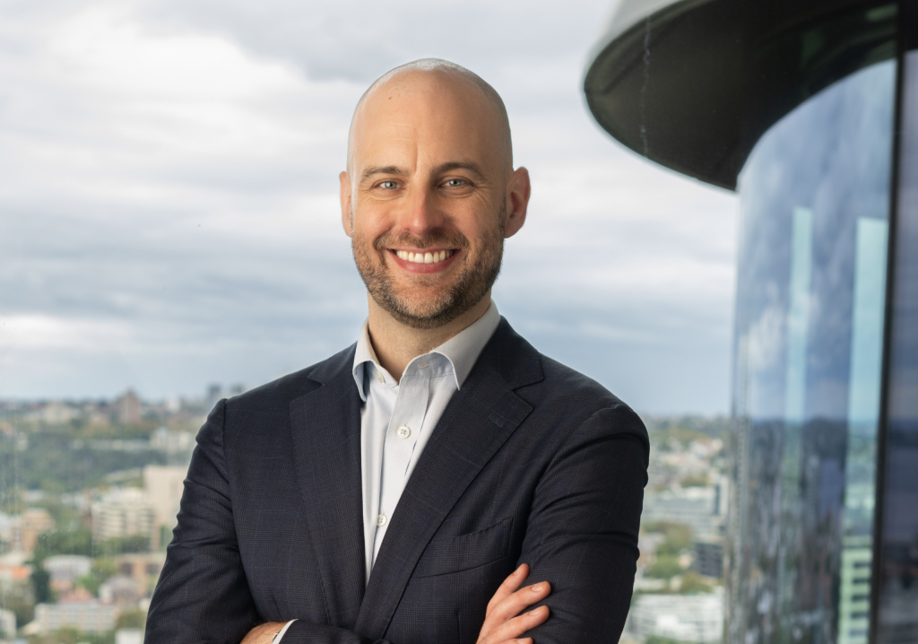 Adam Copp, dressed in a navy suit, smiles at the camera.