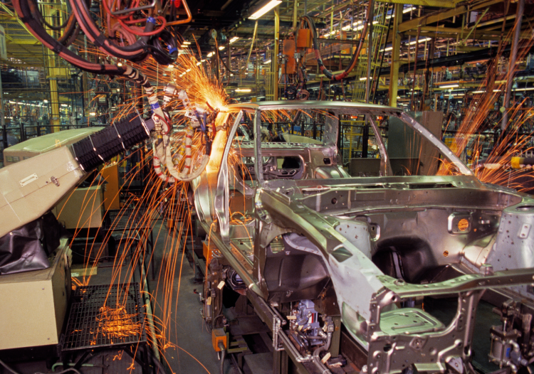 Sparks fly from a car chassis in a busy factory.